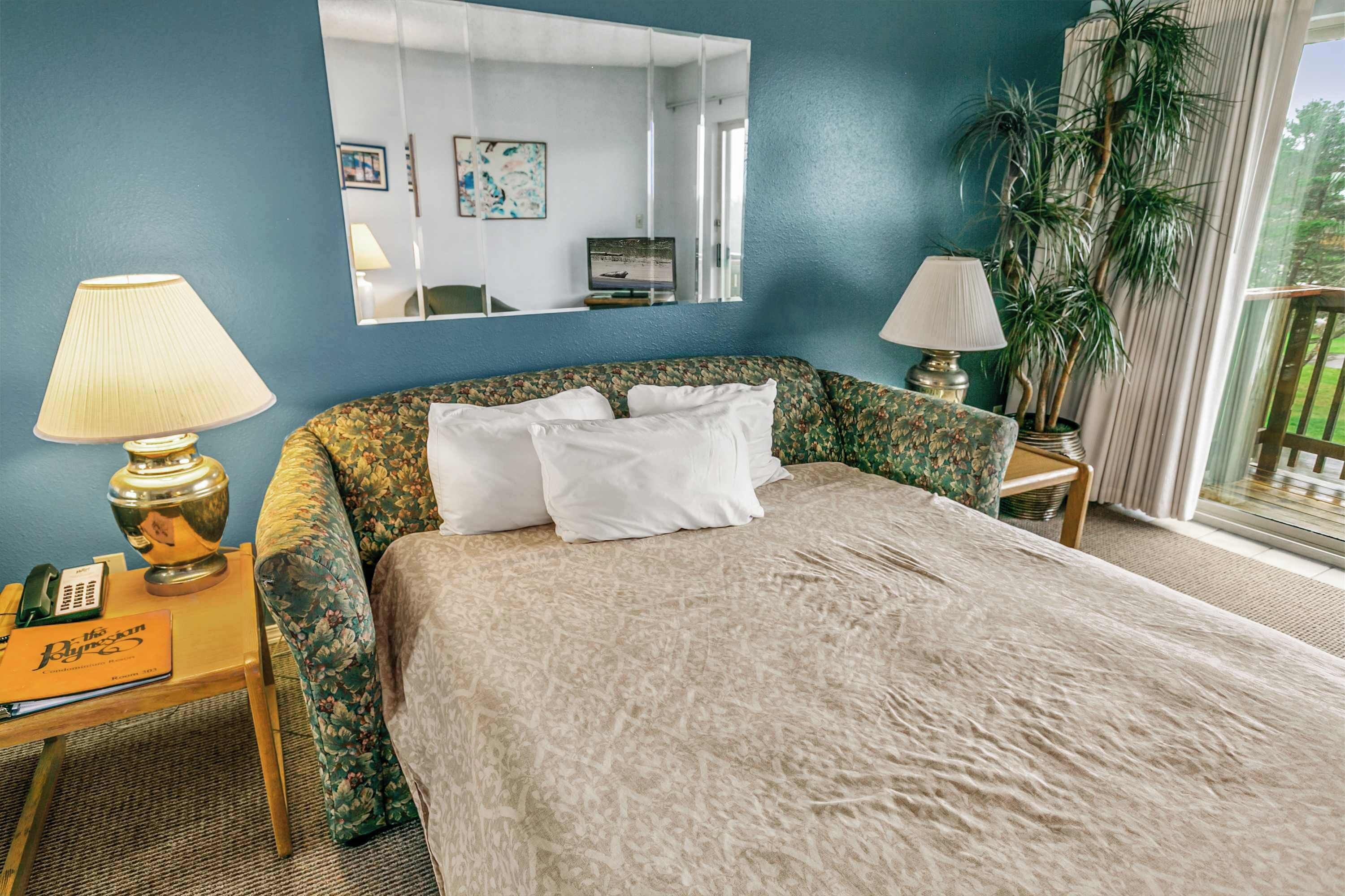 Habitación de hotel con cama, dos mesitas de noche, un espejo, una planta en maceta y vistas al exterior.