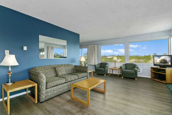 Ein Wohnzimmer mit blauer Wand, einem Holztisch, einem blumigen Sofa, zwei Sesseln, einem Fernseher und einem großen Fenster mit Blick.