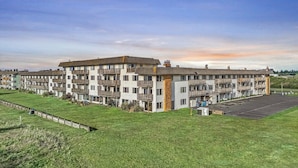 A multi-story hotel with balconies, a parking lot, and a grassy area. - 2-Bedroom Condo - King Bed, Fireplace, & Kitchen! (Ocean Shores)