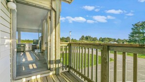 A wooden deck with a railing, a covered porch, and a view of a parking area and greenery. - 2-Bedroom Condo - King Bed, Fireplace, & Kitchen! (Ocean Shores)