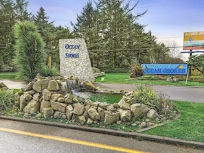 A stone sign with 'Ocean Shores' written on it, a small waterfall feature, and a road with a yellow line. - 2-Bedroom Condo with King Bed and Fireplace - Pool, Hot Tub and Game Room Access! (Ocean Shores)