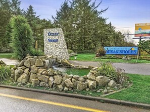 A stone sign with 'Ocean Shores' written on it, a small waterfall feature, and a road with a yellow line. - Ocean View, Pet-Friendly 1-Bedroom Family Suite - Pool and Hot Tub! (Ocean Shores)