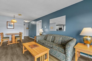 A living room with a sofa, two wooden tables, a dining table with chairs, a hanging light fixture, and a kitchen area visible through an open door. - 2-Bedroom Family Suite With Kitchen - Pet-Friendly (Ocean Shores)