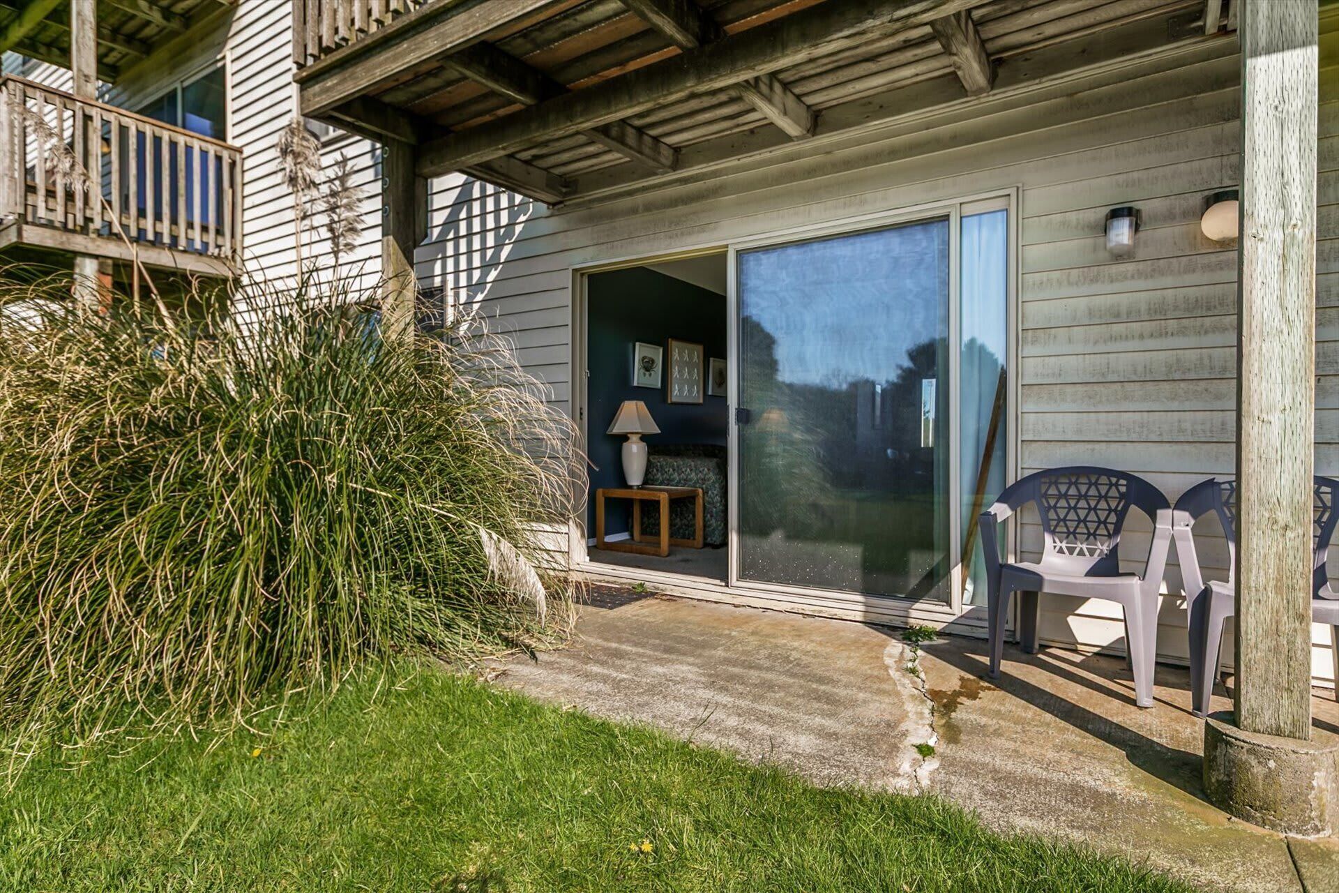 A house with a wooden porch, a sliding glass door, a small table with a lamp, and outdoor chairs.