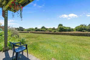 A covered patio with a hanging flower basket, a chair, and a view of a grassy field with a stone wall in the background.