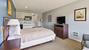 Habitación de hotel con una cama, un escritorio con lámpara, televisión, cómoda y una kitchenette al fondo.