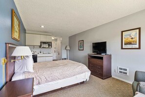 A hotel room with a bed, a desk with a lamp, a television, a dresser, and a kitchenette in the background. - Queen Studio, Ground Floor - Pool & Hot Tub Access (Ocean Shores)