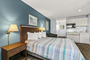 Habitación de hotel con cama, lámparas de noche, escritorio, silla, kitchenette y un cuadro en la pared.