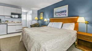 A hotel room with a bed, bedside lamps, a desk, a chair, a painting on the wall, and a kitchenette. - Pet Friendly Condo with King Bed and Kitchenette (Ocean Shores)