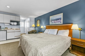 A hotel room with a bed, bedside lamps, a desk, a chair, a painting on the wall, and a kitchenette. - Pet Friendly Condo with King Bed and Kitchenette (Ocean Shores)