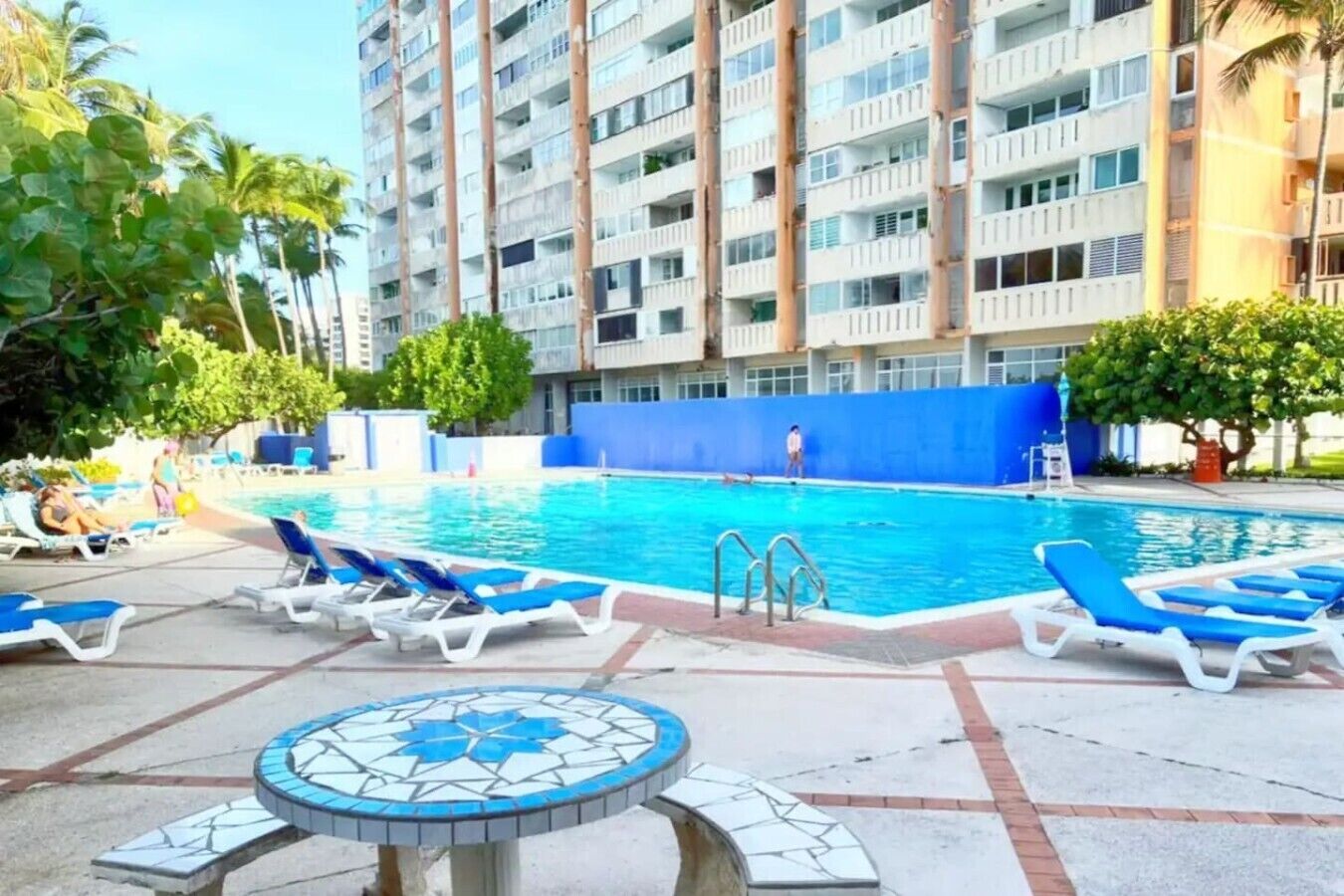 A swimming pool with lounge chairs, a tiled deck, and a high-rise building in the background.