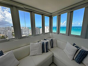 A modern living room with a white sectional sofa, multiple windows offering a view of the ocean and cityscape. - Corner For 2 BEAUTIFUL OCEAN VIEW Pool Beach Parking CONDADO (San Juan)