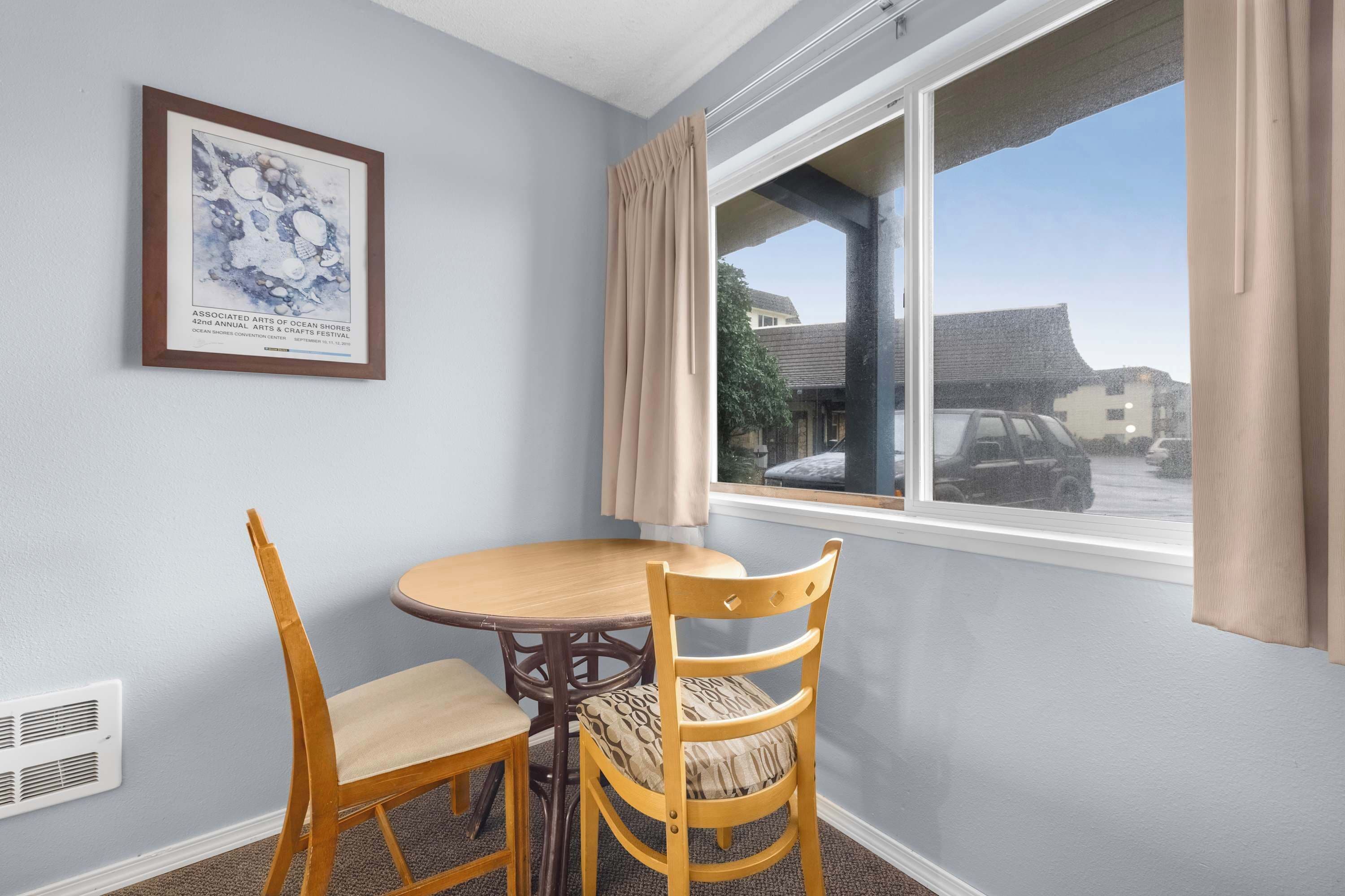 A room with a dining table and chairs, a framed artwork on the wall, and a window with curtains.
