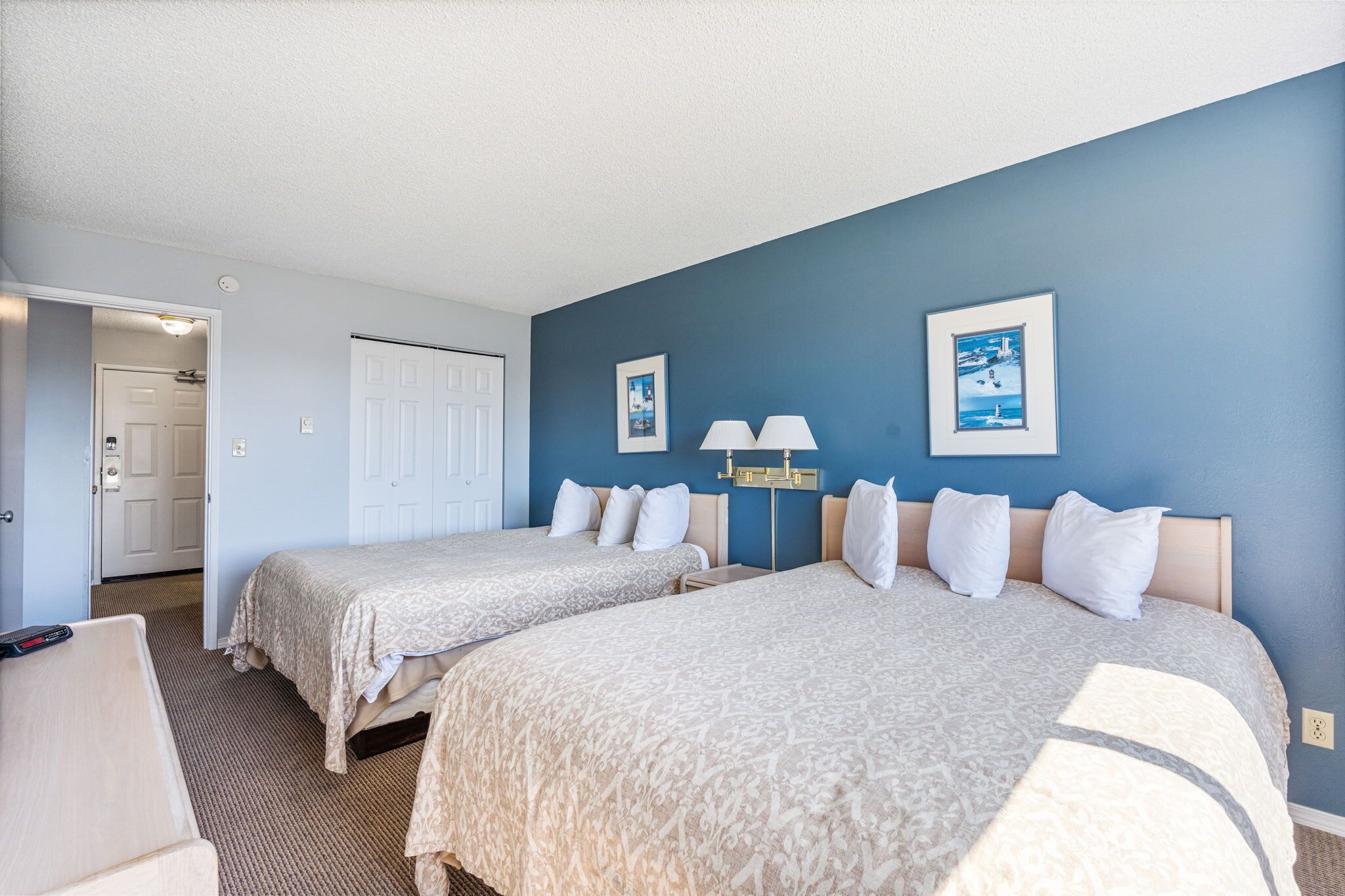 Une chambre d’hôtel avec deux lits, un bureau, une chaise, une porte et un mur orné de deux tableaux encadrés.