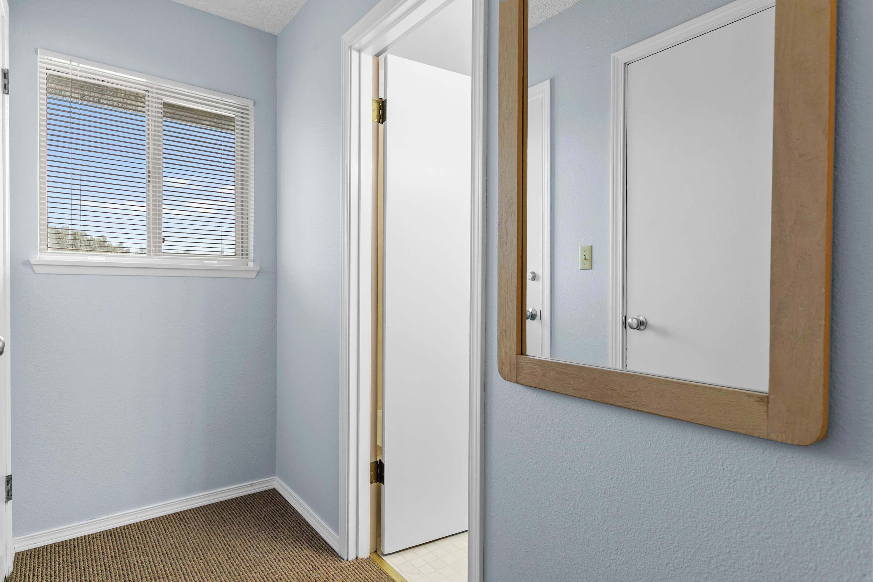 A hallway with a door, a window with blinds, and a mirror on the wall.