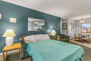 A hotel room with a bed, two bedside tables with lamps, a chair, a desk with chairs, and a view of the outside. - 1-Bedroom Suite For 4 With King Bed & Pool Access (Ocean Shores)