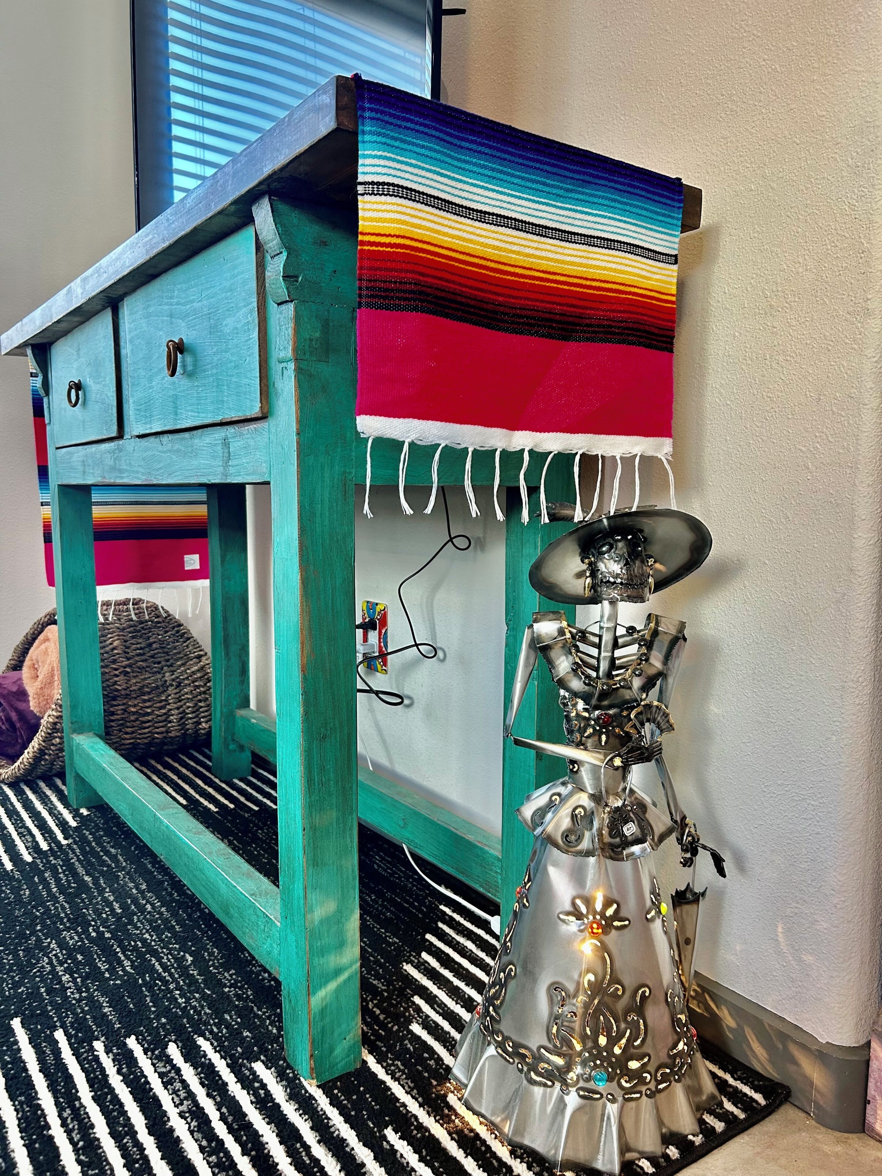 Un bureau de couleur turquoise avec des tiroirs, une couverture colorée en tissu posée dessus, et une sculpture métallique coiffée d’un chapeau.