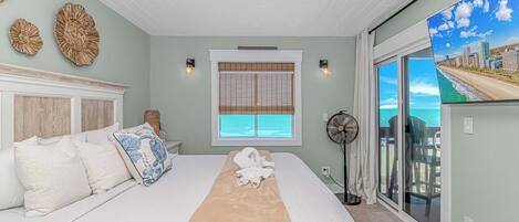 A bedroom with a large bed, a window with blinds, a fan, and a view of the ocean.