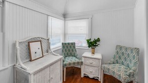 A room with white paneled walls, a ceiling fan, a wicker bench with a picture frame, two patterned chairs, and a small white side table with a plant. - 4BR Beach Block Home w/ Washer-Dryer, Free Parking (Seaside Heights)
