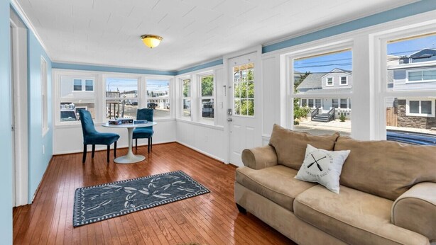 A room with a wooden floor, a beige sofa, a blue chair, a table with a rug, and large windows offering a view of houses. - 4BR Beach Block Home w/ Washer-Dryer, Free Parking (Seaside Heights)