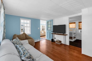 Ein Zimmer mit einer Couch, einem Fernseher auf einem Schrank und einer Tür, die auf einen Balkon führt, mit Blick ins Freie.