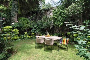 Um jardim com mesa e cadeiras, rodeado por vegetação exuberante e um muro de pedra.