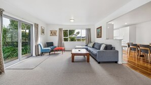 A spacious living room with a grey sofa, a coffee table, and a dining area with wooden chairs. - Sundown on Cologne - Martinborough Holiday Home (Martinborough)