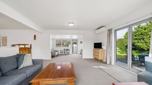 A modern living room with a sofa, coffee table, TV, and a view of a garden through large windows. - Sundown on Cologne - Martinborough Holiday Home (Martinborough)