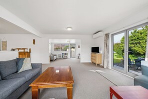 Un salon moderne avec un canapé, une table basse, un téléviseur et une vue sur un jardin grâce à de grandes fenêtres.