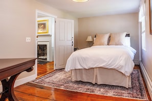 A bedroom with a bed, a nightstand, a lamp, a wooden floor, and a door leading to another room.
