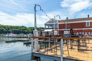 A waterfront restaurant with outdoor seating, a dock, and boats in the background.