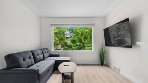 A modern living room with a grey sectional sofa, a small round table, a window with a view of greenery, and a mounted TV. - 2 Bedroom + Pull out Sofa | Min to UofC (Calgary)