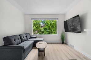 A modern living room with a grey sectional sofa, a small round table, a window with a view of greenery, and a mounted TV. - 2 Bedroom + Pull out Sofa | Min to UofC (Calgary)