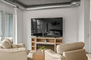 A living room with a large flat-screen TV on a wooden stand, two beige sofas, a small coffee table, and a fireplace. - `The Cedar` | Mins to Stampede | Patio Fire-pit (Calgary)