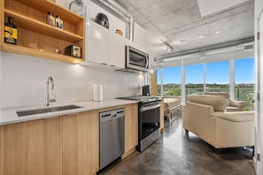 A modern kitchen with a dishwasher, oven, and microwave, wooden shelves, and a view of the outdoors. - `The Cedar` | Mins to Stampede | Patio Fire-pit (Calgary)