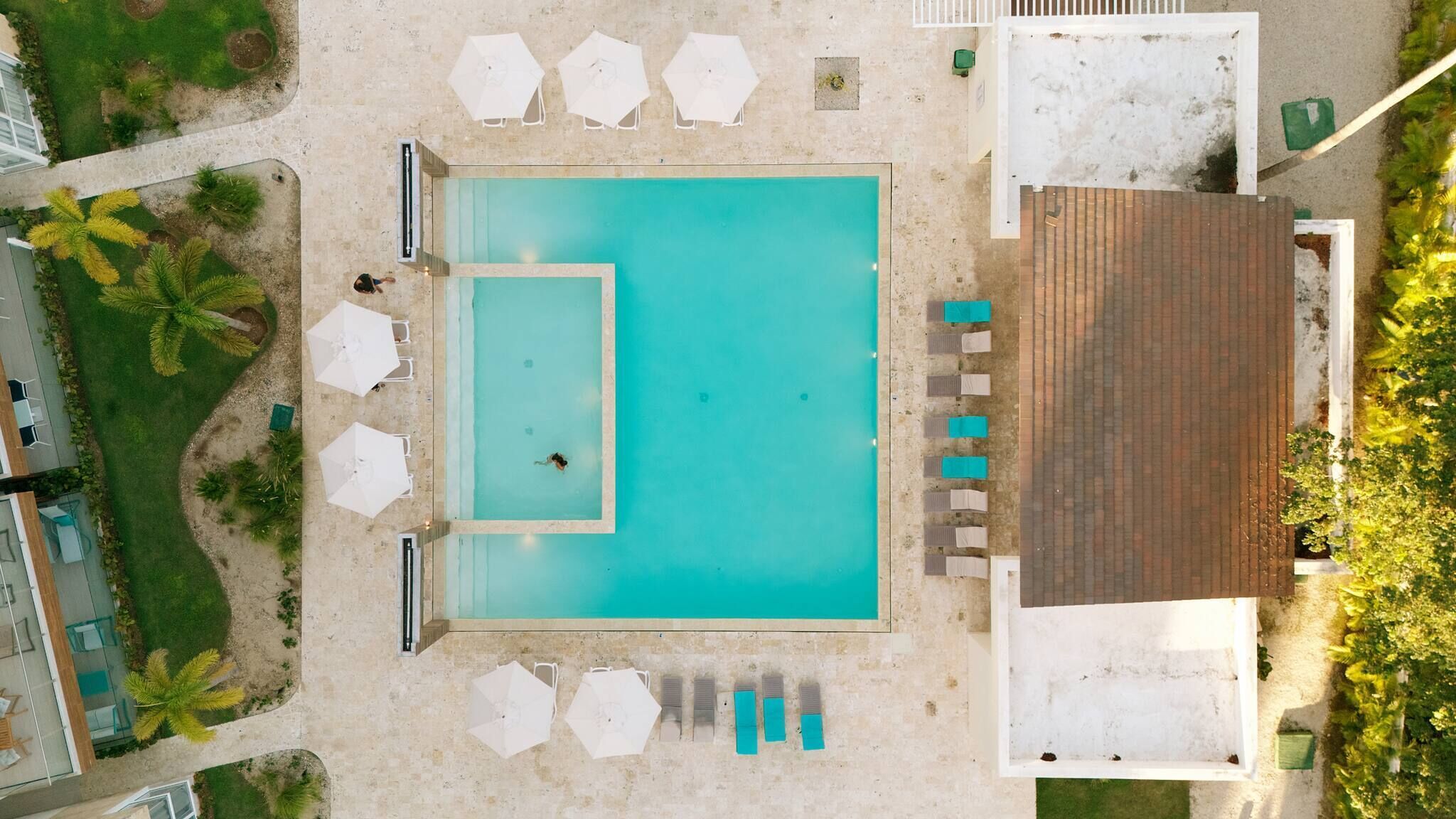 An aerial view of a swimming pool with a smaller inner pool, surrounded by lounge chairs and umbrellas, with a building and palm trees nearby.