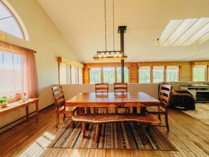 A dining area with a wooden table and chairs, a bench, a hanging light fixture, and large windows with a view of trees. - Sunrise Ridge•SwimSpa•Pikes Peak Views • Sleeps 10 (Divide)