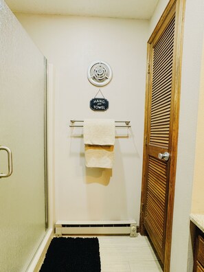 A bathroom with a towel rack, a sign that says 'HANG your towel,' a radiator, a wooden door, and a black rug. - Sunrise Ridge•SwimSpa•Pikes Peak Views • Sleeps 10 (Divide)