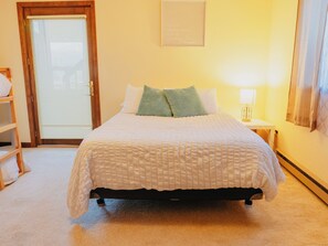 A neatly made bed with a white comforter and pillows, a bedside lamp, a wooden nightstand, a door with a window, and a wall-mounted sign. - Sunrise Ridge•SwimSpa•Pikes Peak Views • Sleeps 10 (Divide)