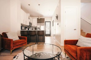 A modern living room with a glass coffee table, two sofas, and a kitchen in the background.