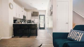 Een smalle gang met aan één kant een keuken, een eethoek met een tafel en stoelen, en een woonkamer met een bank en een kussen met 'LAUGH' erop.