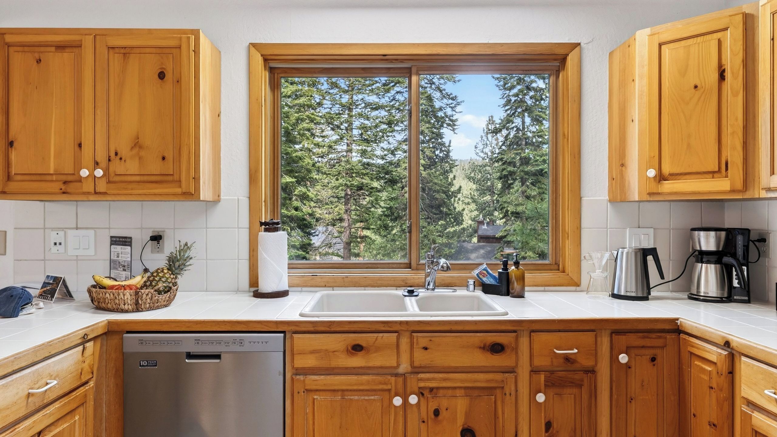 A kitchen with wooden cabinets, a stainless steel dishwasher, a window with a view of trees, and a coffee maker.