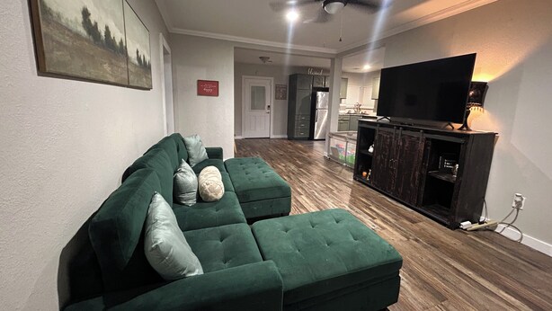 A living room with a green sectional sofa, a flat-screen TV on a wooden stand, a ceiling fan, and a kitchen area in the background. - Comfy Crossroads - 1mi to HWY & 4mi to Downtown (Wake Village)