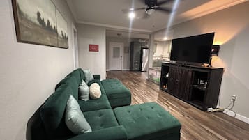 Un salon avec un canapé d’angle vert, un téléviseur à écran plat posé sur un meuble en bois, un ventilateur de plafond et, au fond, un coin cuisine.