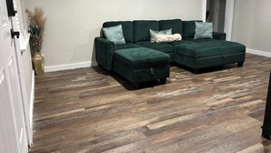 A living room with a sectional sofa, a wooden herringbone floor, a vase with dried plants, and two landscape paintings on the wall. - Comfy Crossroads - 1mi to HWY & 4mi to Downtown (Wake Village)