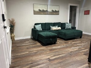 Una sala de estar con un sofá seccional, piso de madera en patrón espiga, un jarrón con plantas secas y dos cuadros de paisajes en la pared.