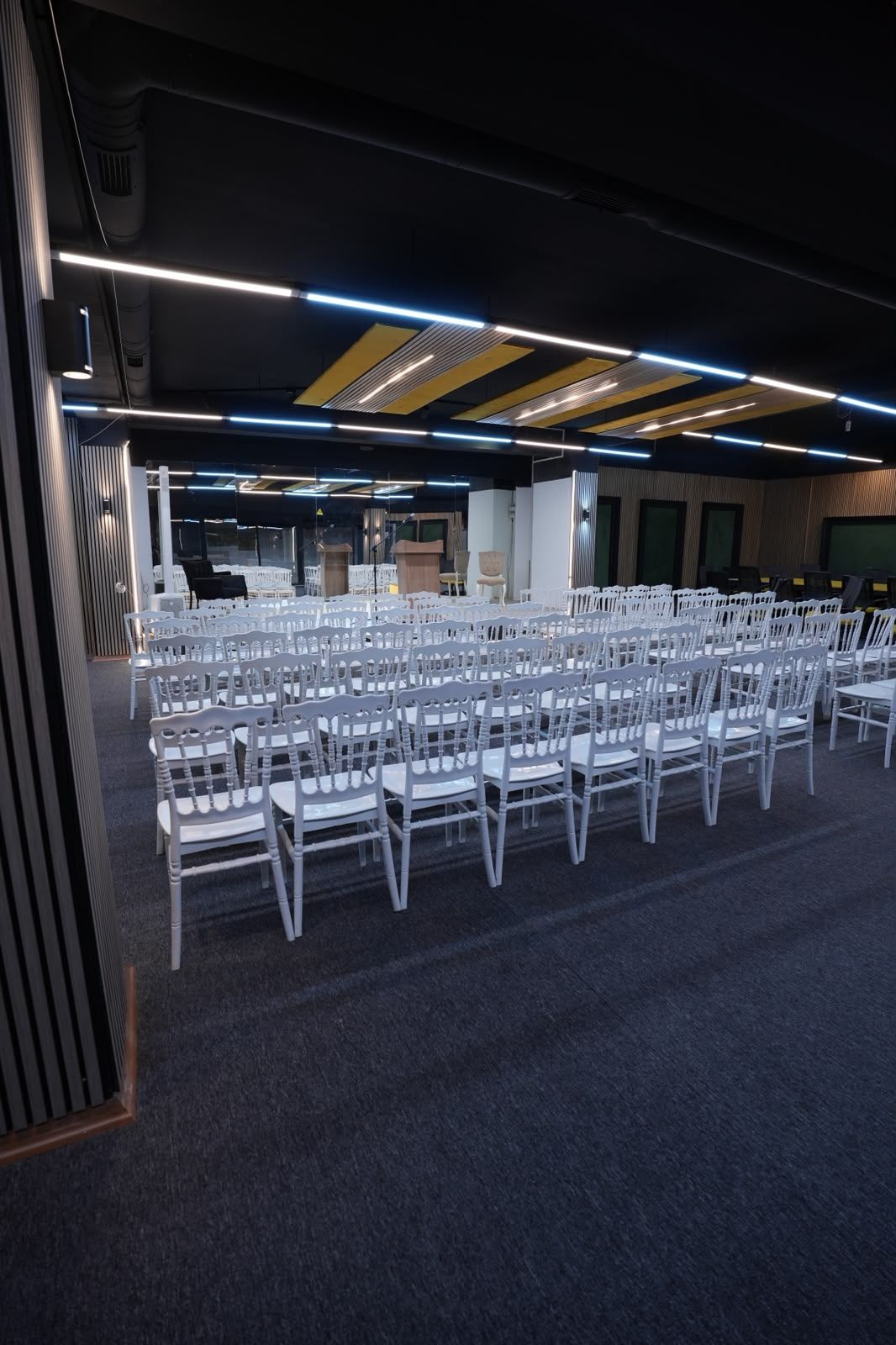 Uma sala de conferência com fileiras de cadeiras brancas, um palco com um pódio e piso coberto de carpete.