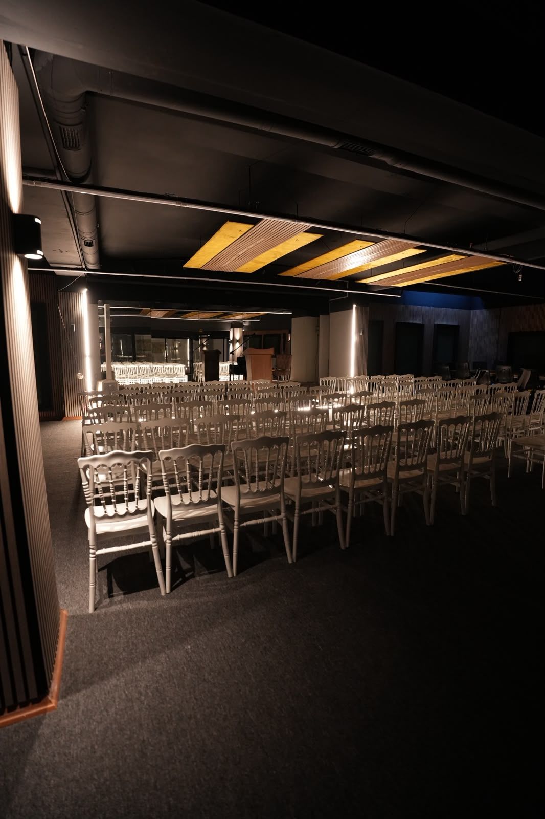 A conference room with rows of chairs and tables, a stage with a podium, and a ceiling with linear light fixtures.