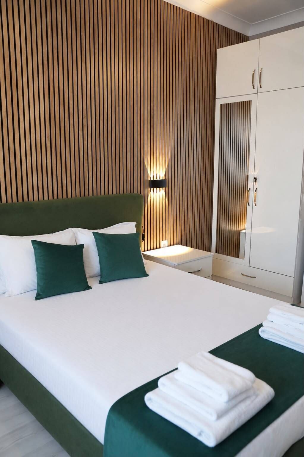 Ein Hotelzimmer mit einem Bett, grünen Kissen, einem Nachttisch, einer Wandleuchte und einem Kleiderschrank.