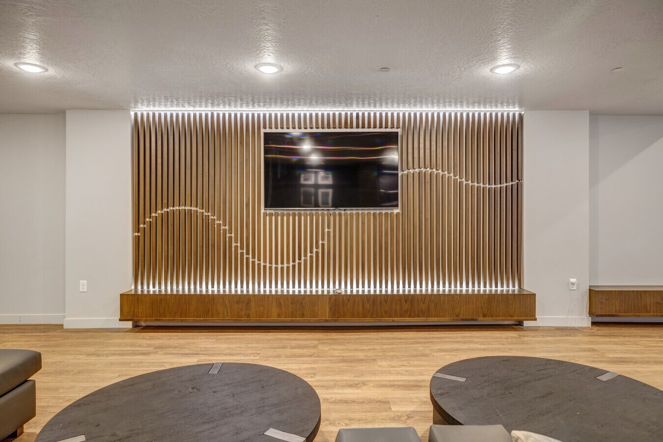 A modern waiting area with a large flat-screen TV mounted on a wooden panel wall, two round tables, and a ceiling with recessed lighting.